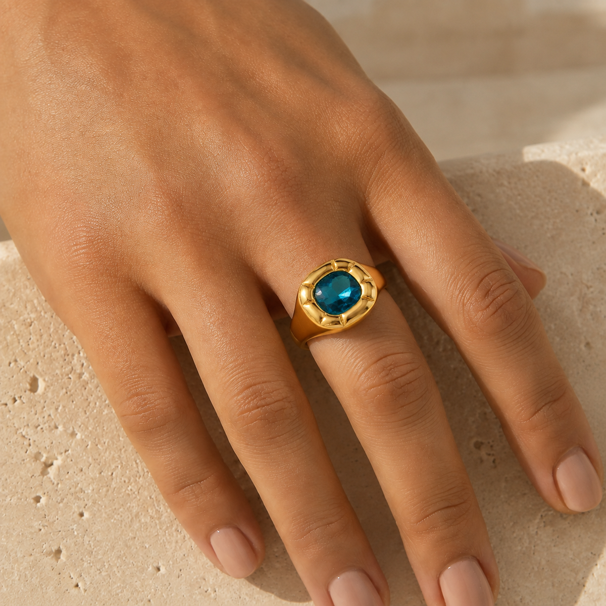 Paloma Cushion Stone Ring – gold ring with a blue stone worn on a hand resting on stone