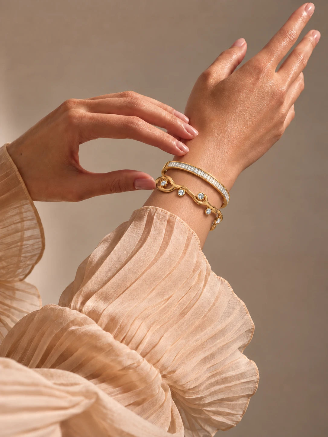 Two gold TAIA bracelets with clear stones, styled with a cream blouse